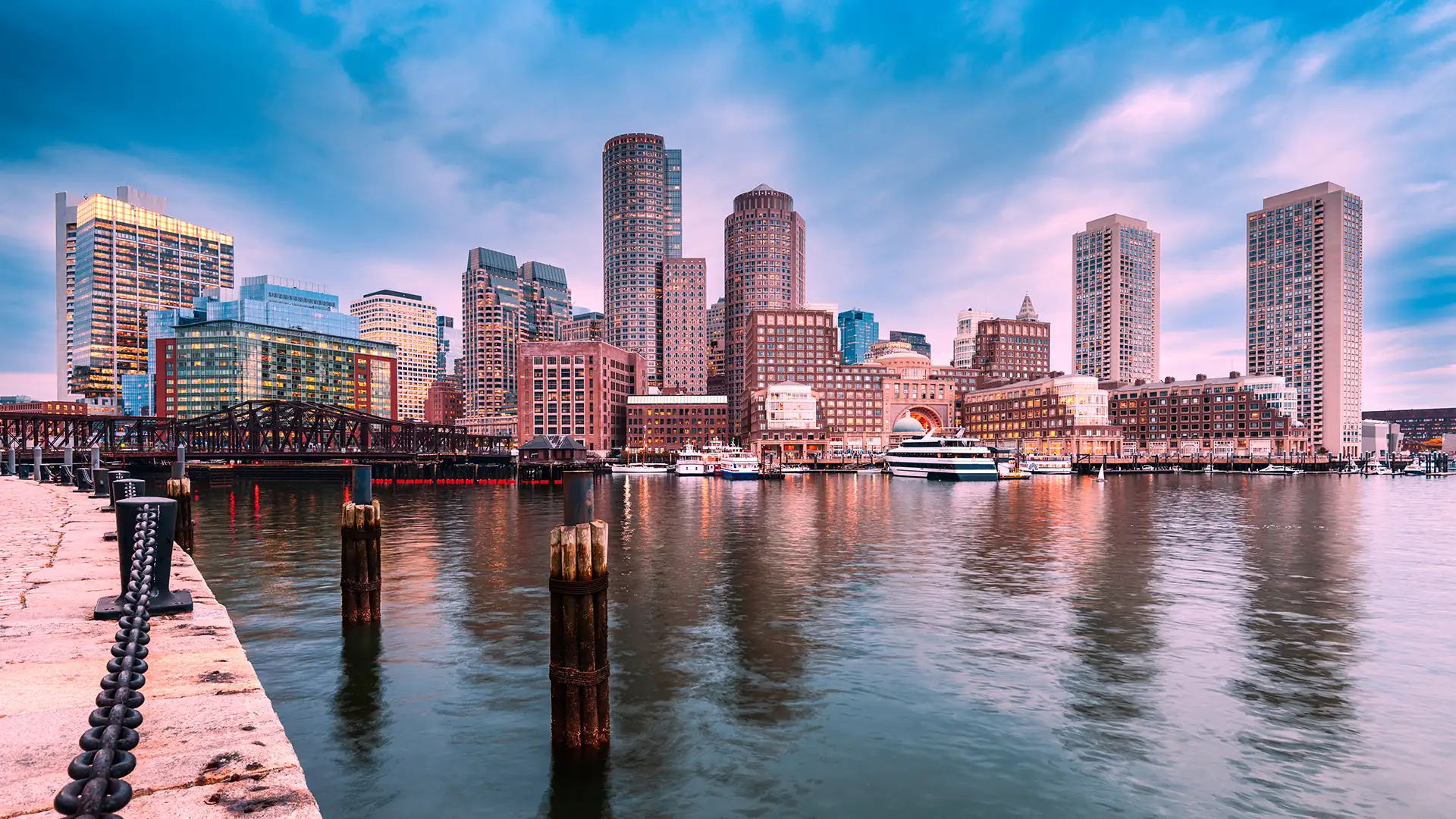 Boston Harbor and the city skyline at sunset, seen from a wharf.